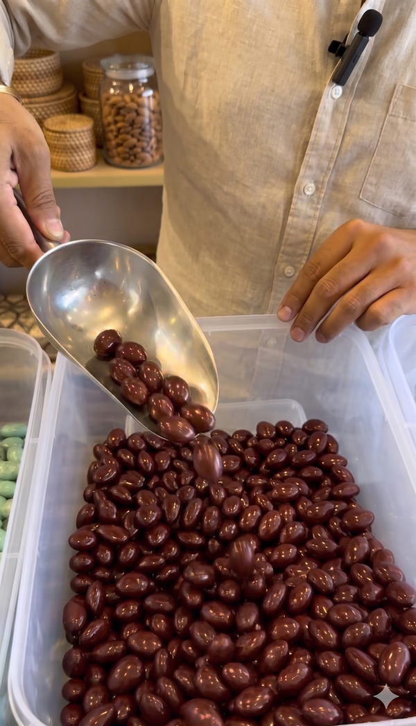 Person scooping chocolate-covered beans from a container with a metal scoop.